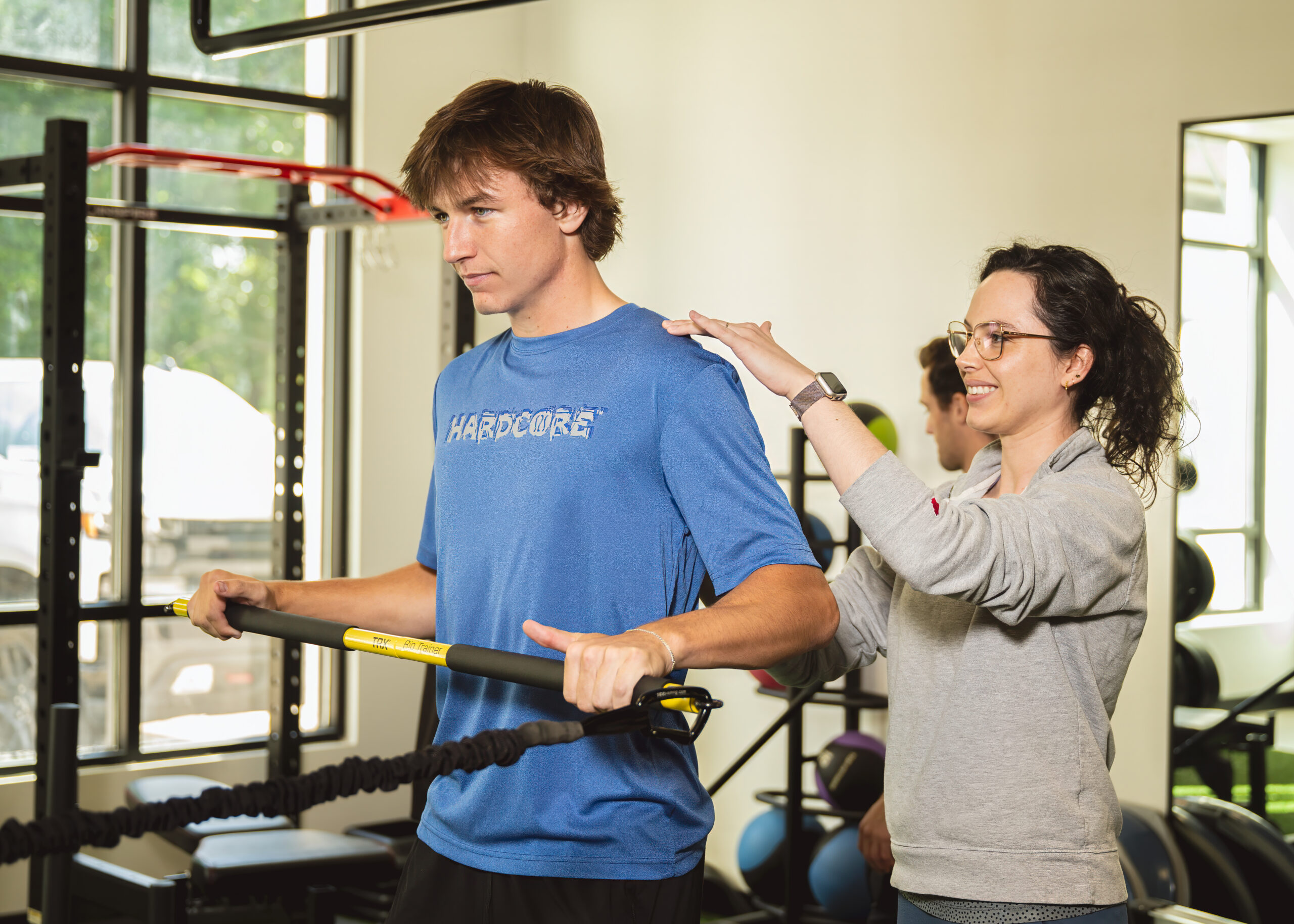 Young male athlete engaged in sports physical therapy, assisted by a female therapist, using resistance bands in a fitness center setting.