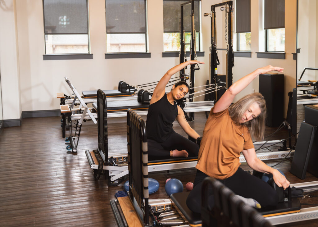Two women performing controlled mobility exercises on reformer machines in a physical therapy studio, focusing on scapular stabilization and improving posture to alleviate neck pain.